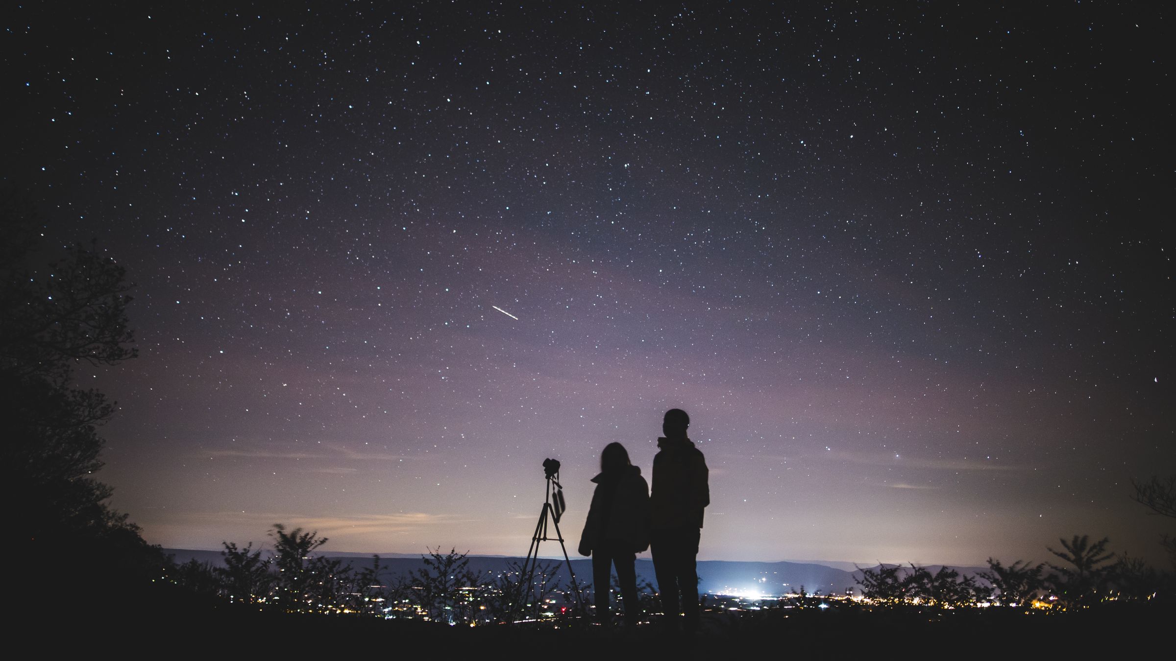 Two stargazers and a telescope under a glowing city sky