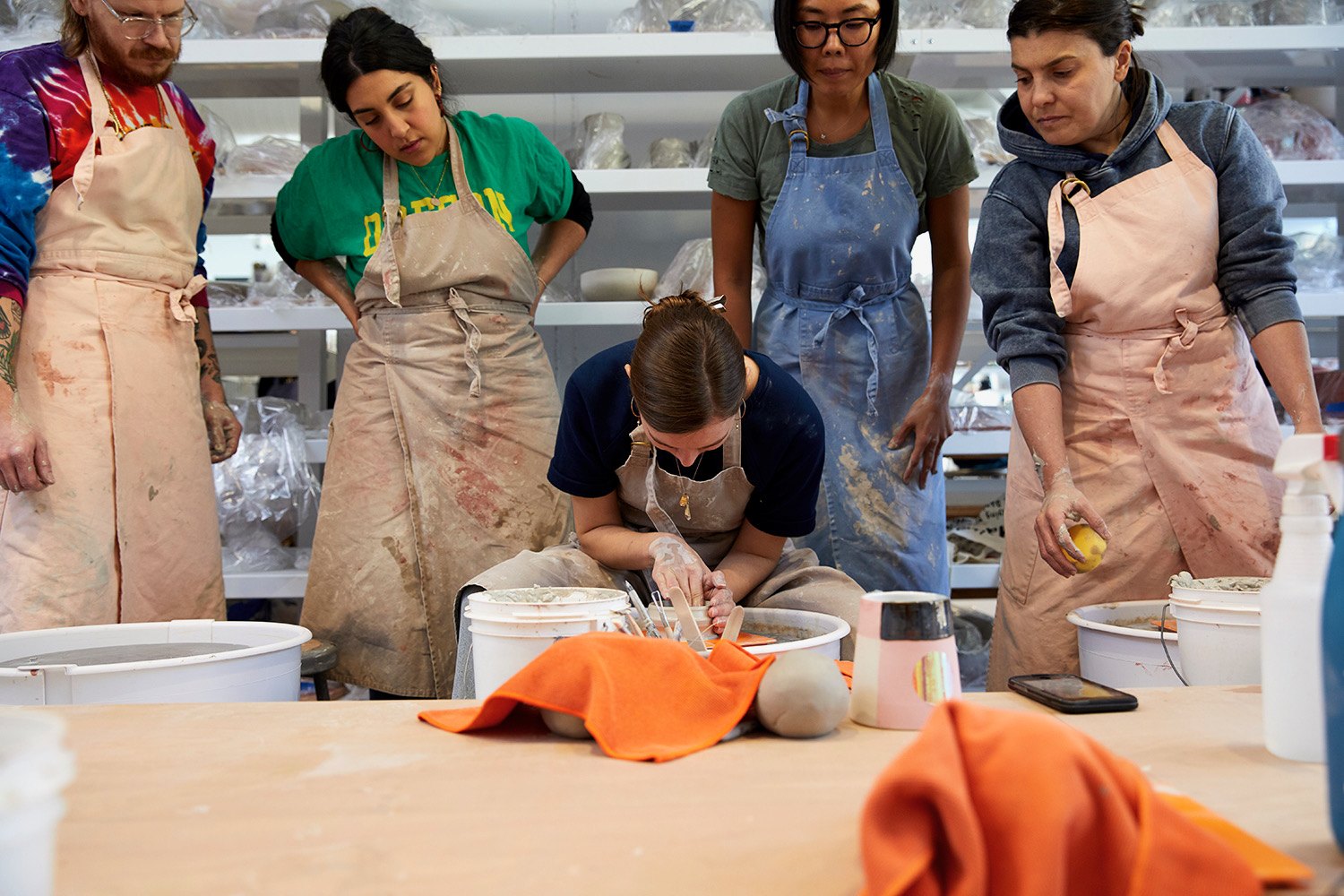 Students in aprons gather round as an instructor centers clay on the wheel at BKLYN CLAY