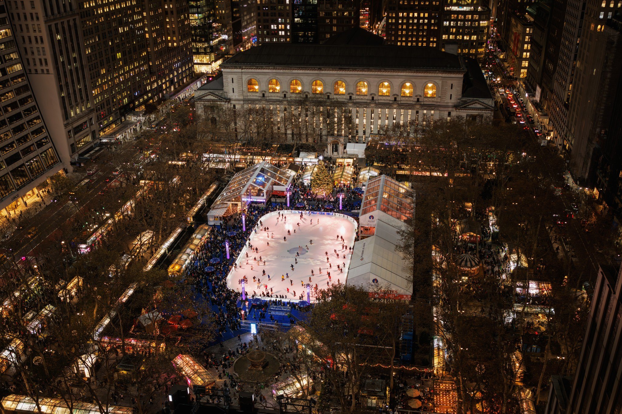 Skaters circling the rink at Bryant Park's Winter Village beneath Midtown towers, snowflake umbrellas in the foreground