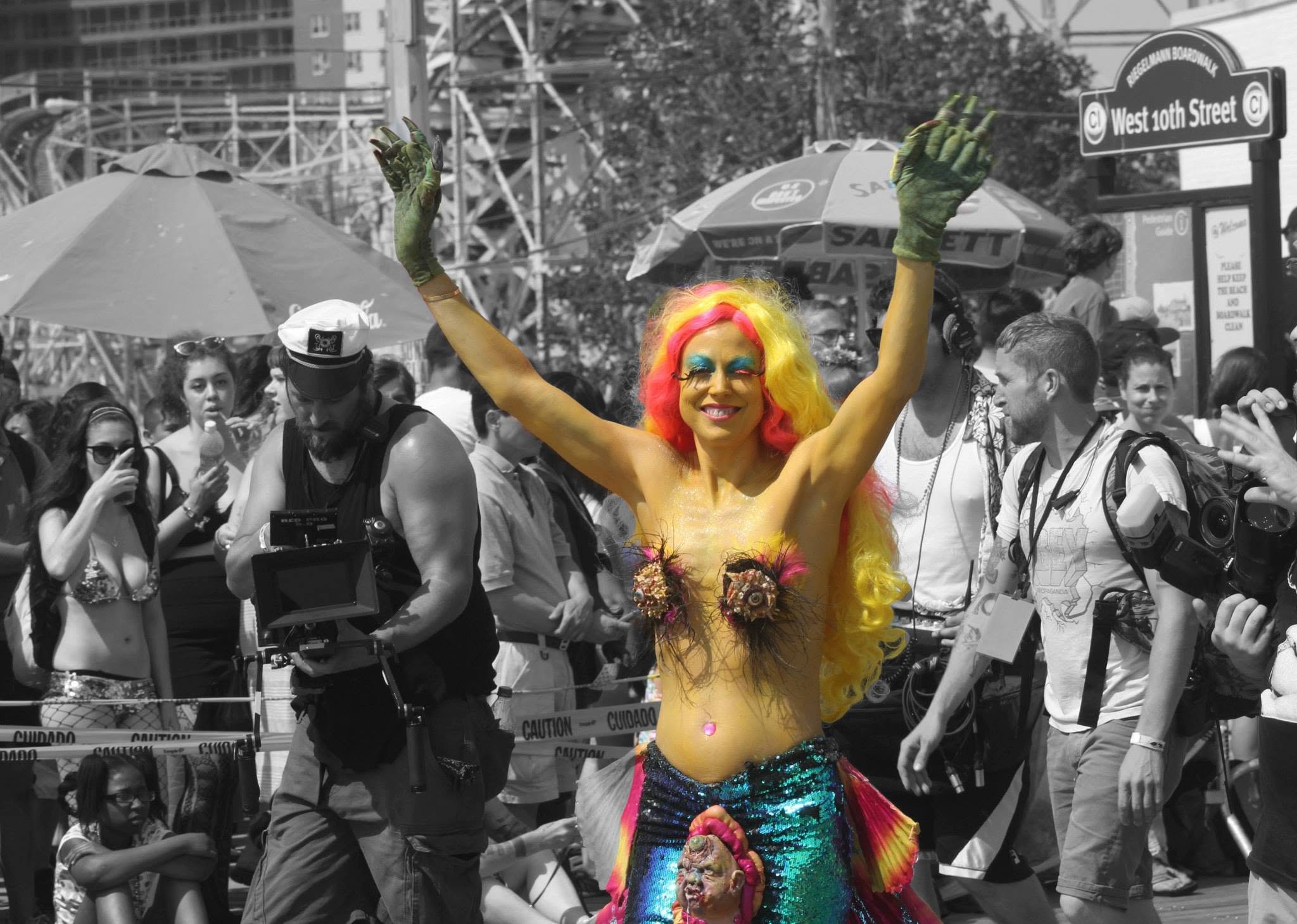 A mermaid in a rainbow wig and sequined tail throws her arms up on the Coney Island boardwalk, the crowd behind her in black and white