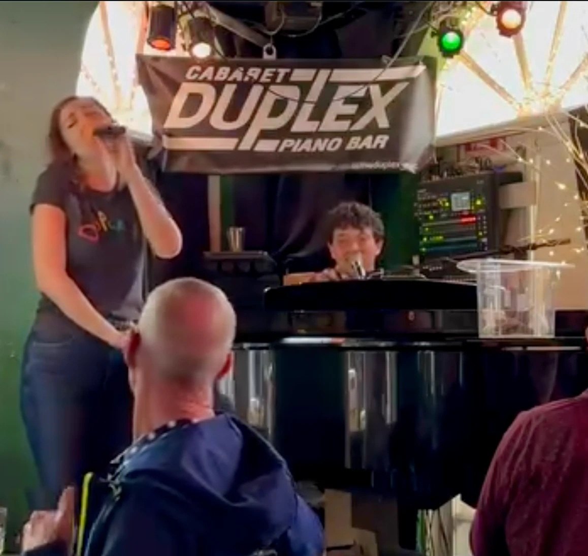 A singer and the pianist at the grand piano under the 'Cabaret Duplex Piano Bar' banner, regulars gathered round