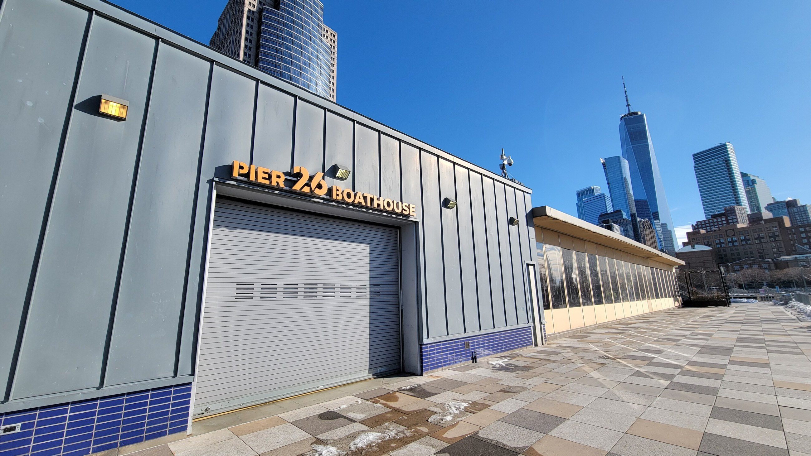 The Pier 26 Boathouse in Hudson River Park, its roll-up kayak bay doors under the 'Pier 26 Boathouse' sign, with One World Trade Center behind