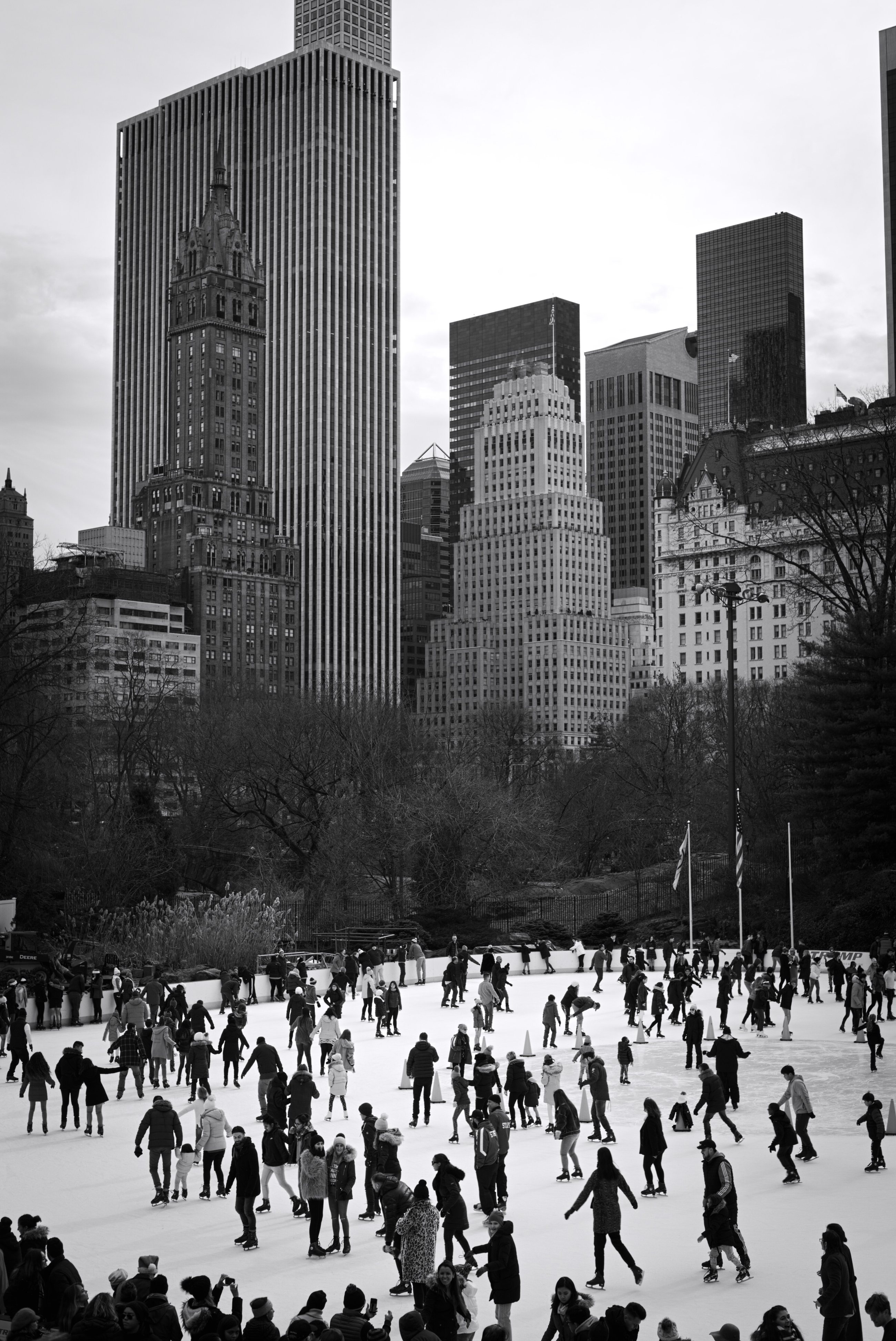 Black-and-white view of skaters on Wollman Rink in Central Park, with the Midtown skyline including the Sherry-Netherland rising behind the trees