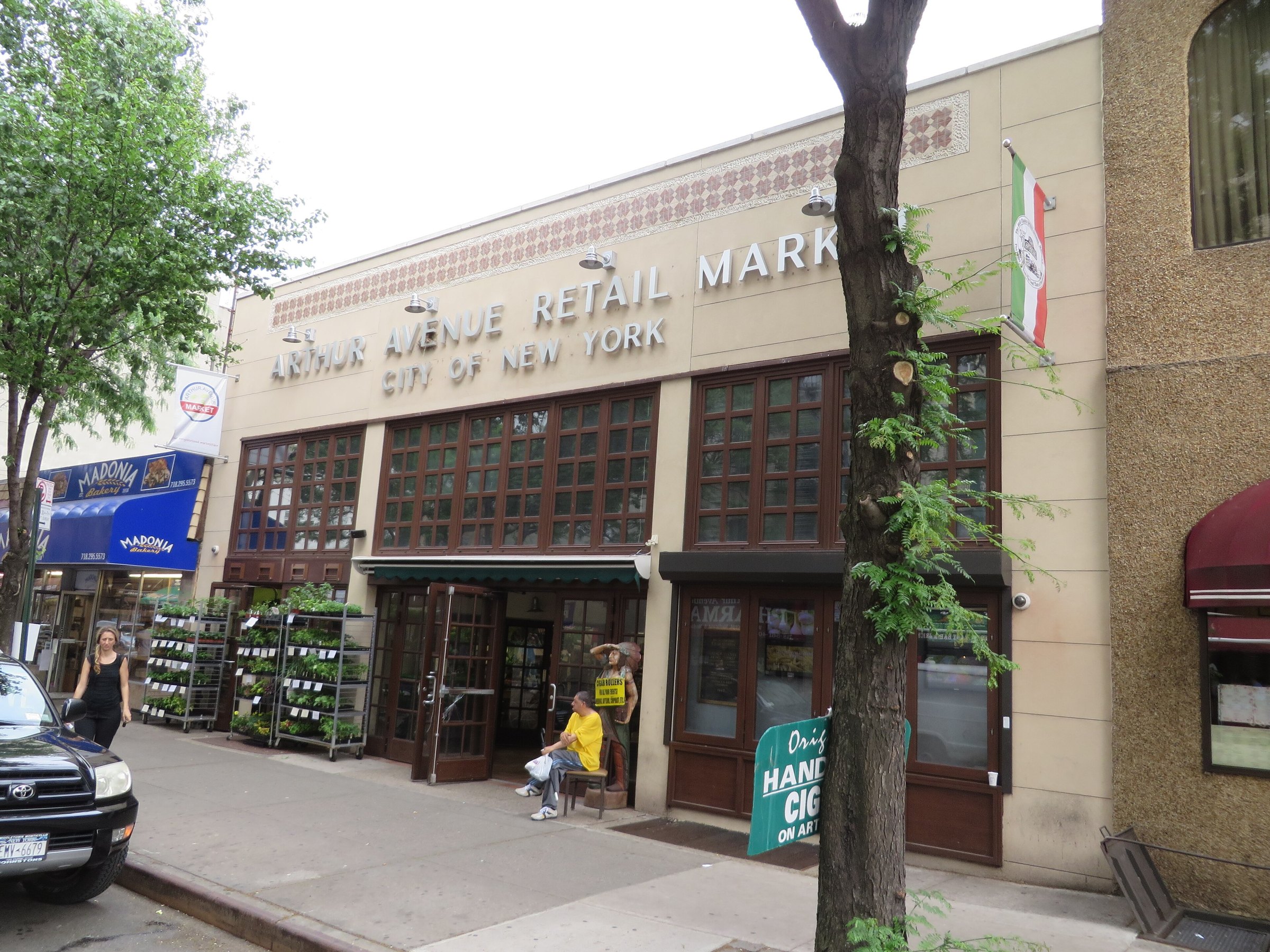 The Arthur Avenue Retail Market's front and sign on a sunny day in the Bronx, with produce stacked by the door