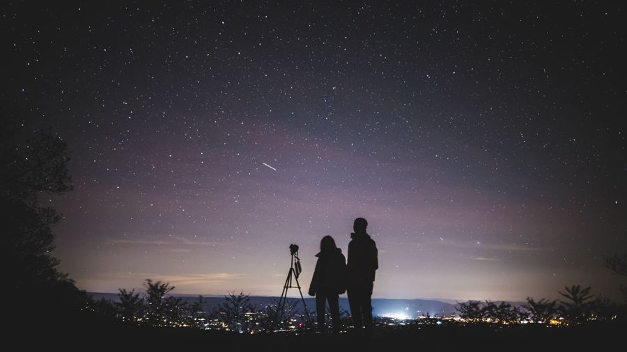 Two stargazers and a telescope under a glowing city sky