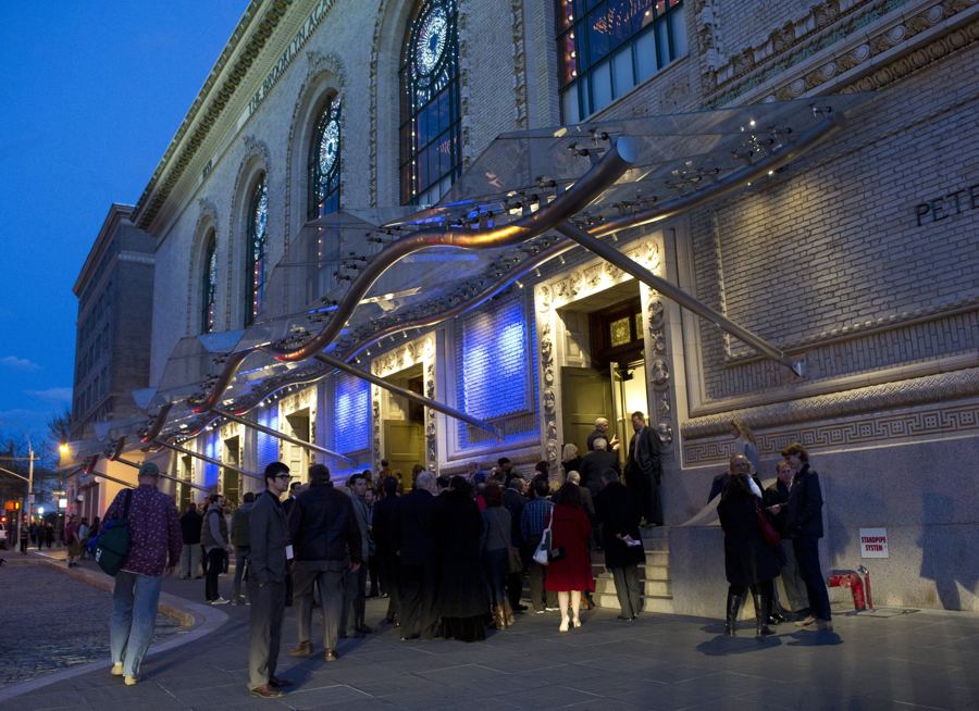 An audience gathering under the glass canopy of the Opera House entrance on an evening at BAM