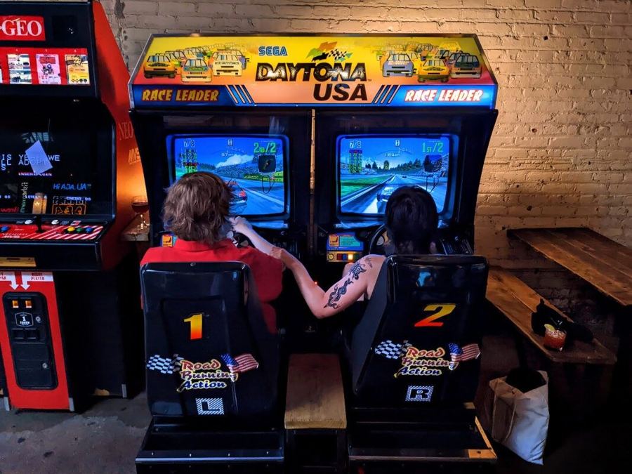 Two players side by side in the Daytona USA racing seats at Barcade in Williamsburg, arcade cabinets along the brick wall