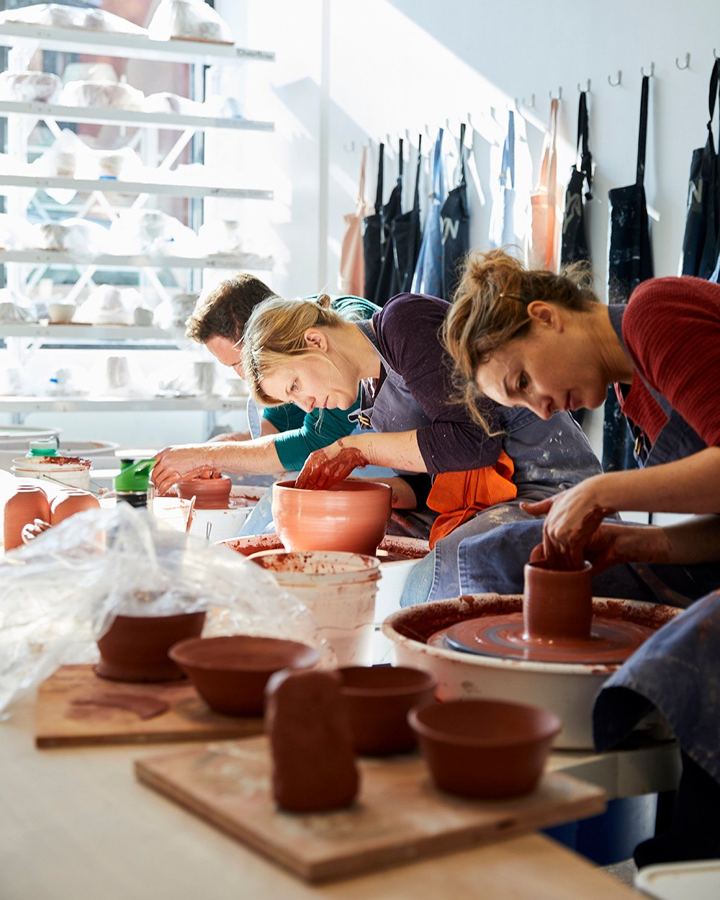 A row of students throwing red clay bowls on pottery wheels in BKLYN CLAY's bright studio