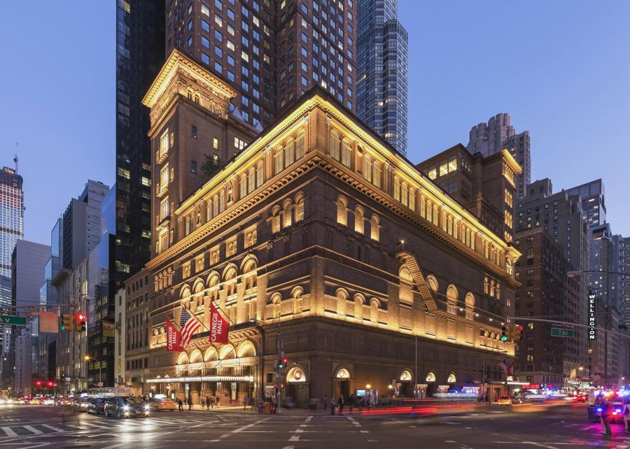 Carnegie Hall's brick and terracotta facade lit gold at dusk on the corner of 57th Street and Seventh Avenue, flags over the entrance