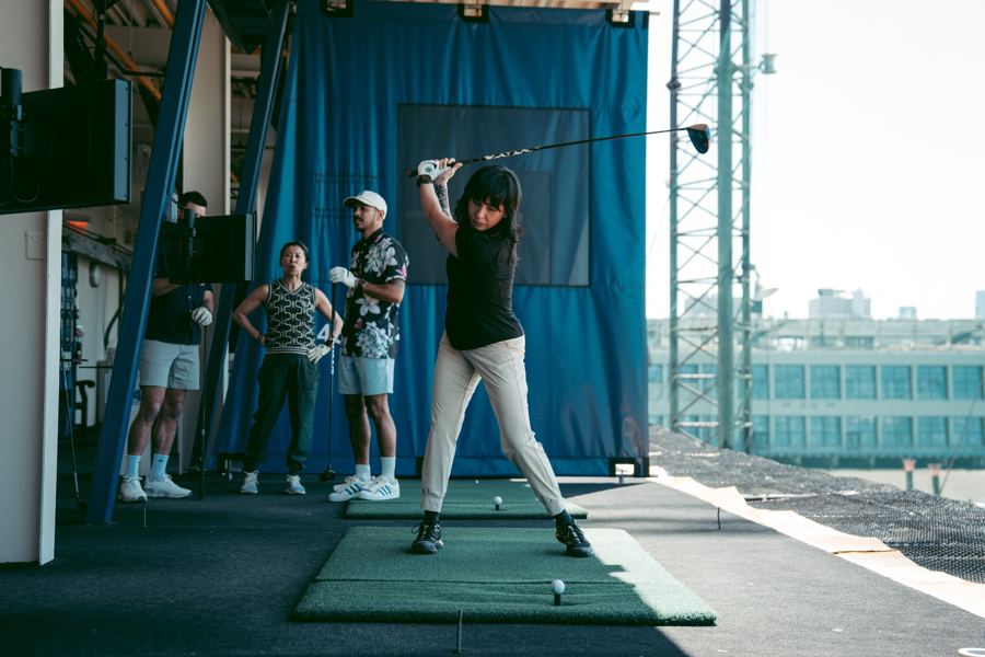 A golfer mid-swing in a hitting bay while friends watch at Chelsea Piers Golf Club