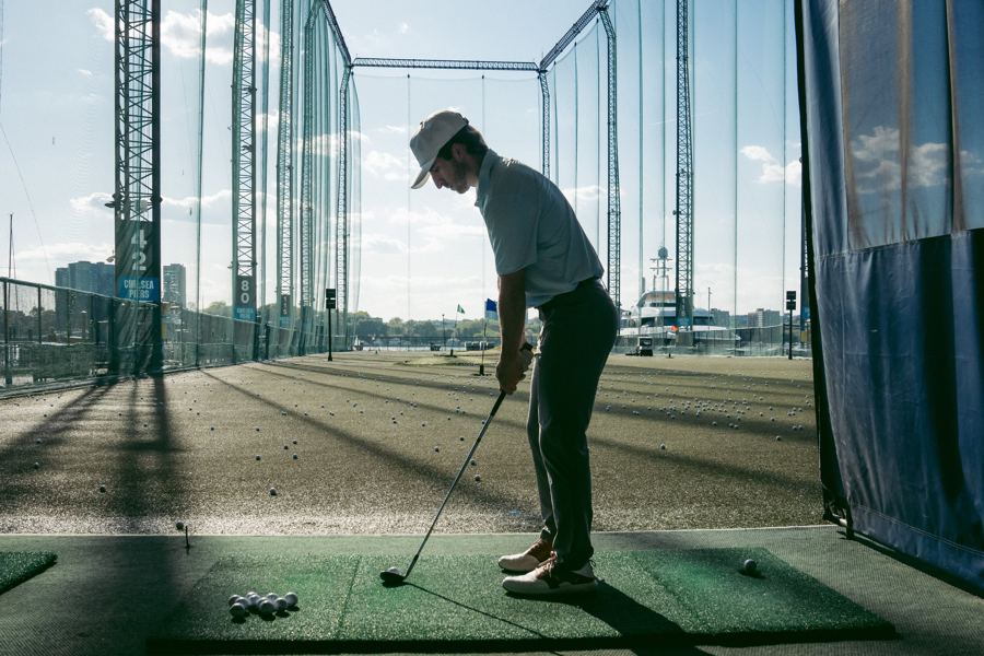 A golfer lining up a shot from a bay, a tray of balls at his feet and the range stretching toward the river