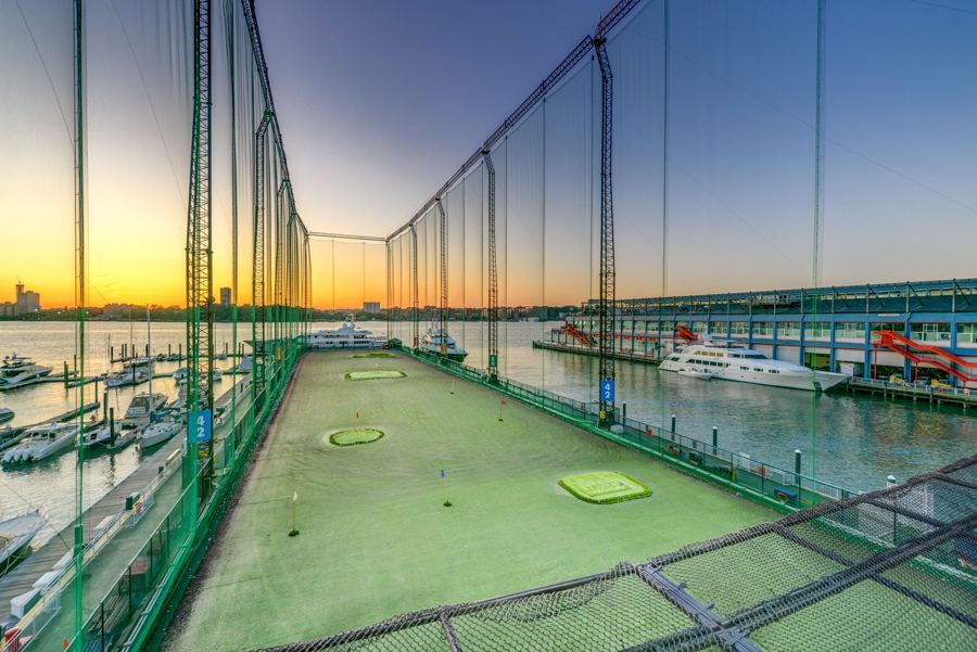 The netted driving range at Chelsea Piers Golf Club at sunset, target greens on the turf and the Hudson marina alongside