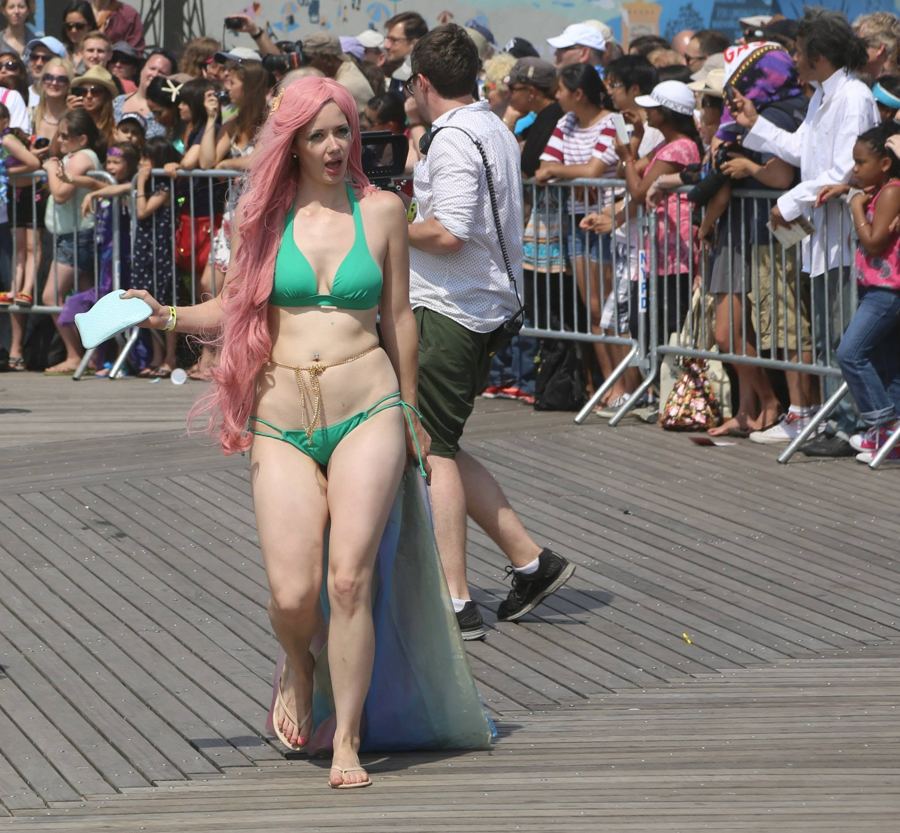 A pink-haired mermaid in a green shell top walks the Coney Island boardwalk past the parade crowd