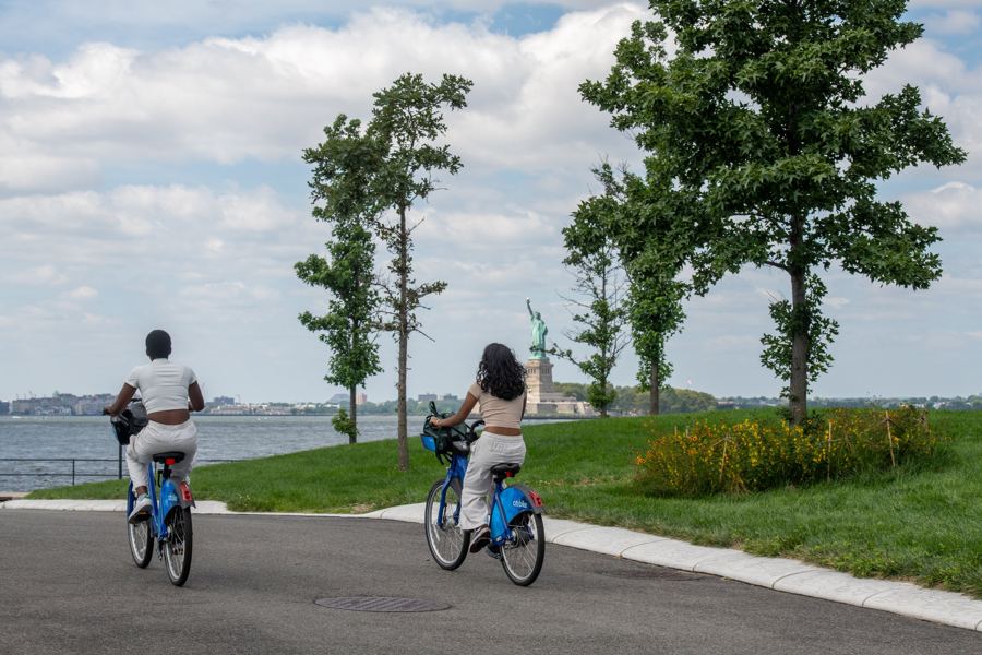Two riders on Citi Bikes along a path through The Hills, the Statue of Liberty across the water