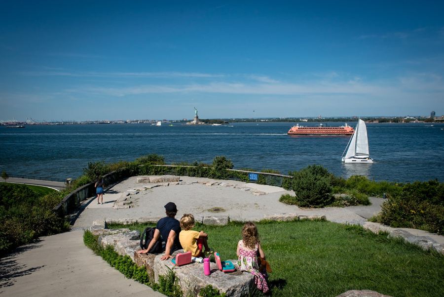 Visitors resting at the top of Outlook Hill on Governors Island, the harbor, a Staten Island ferry and the Statue of Liberty beyond
