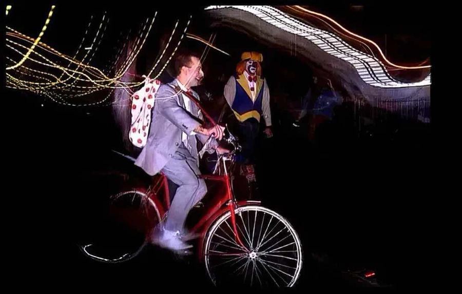 A Pee-wee Herman costume on a red bicycle, streaked with parade lights