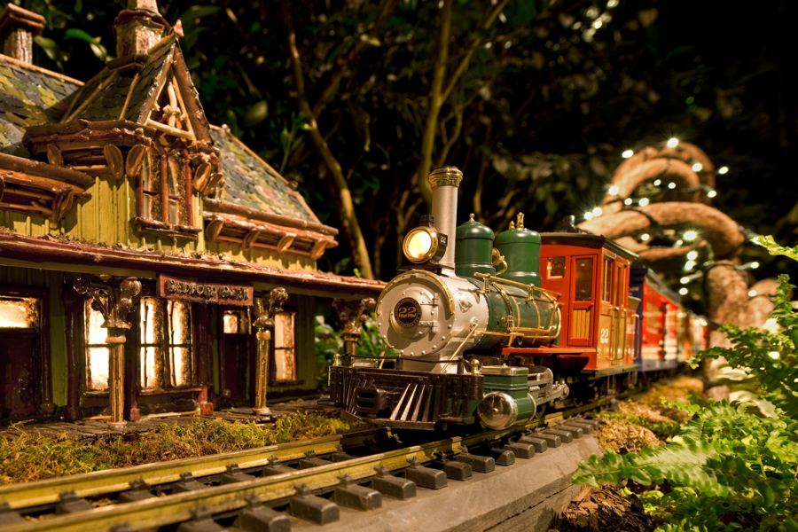 A model steam train and red coach pulling past a plant-made station at the Holiday Train Show
