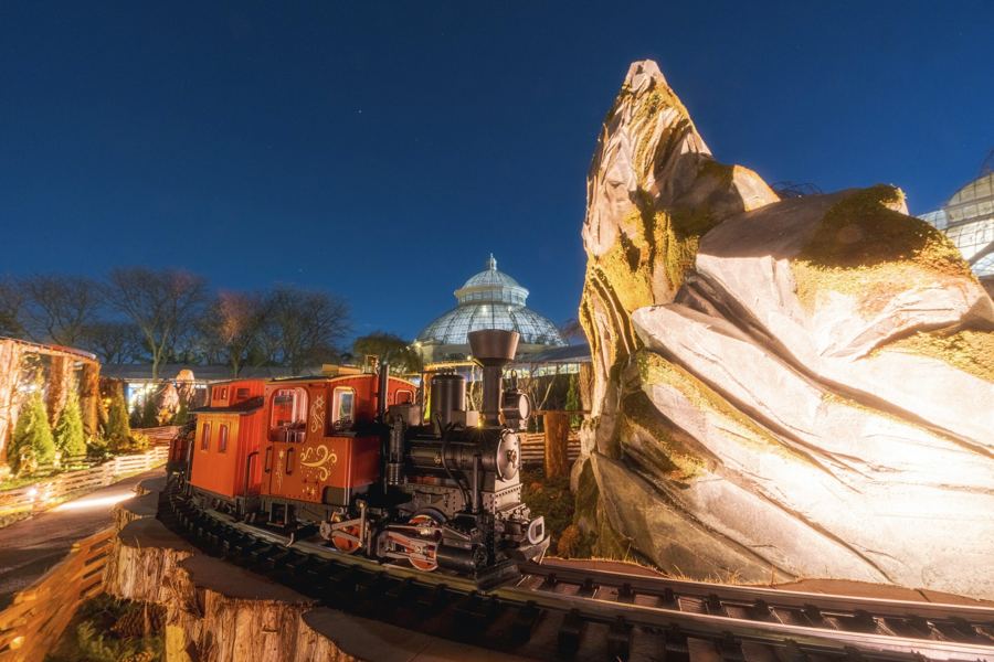 A model train winding through the outdoor woodland expansion at dusk, the conservatory lit behind