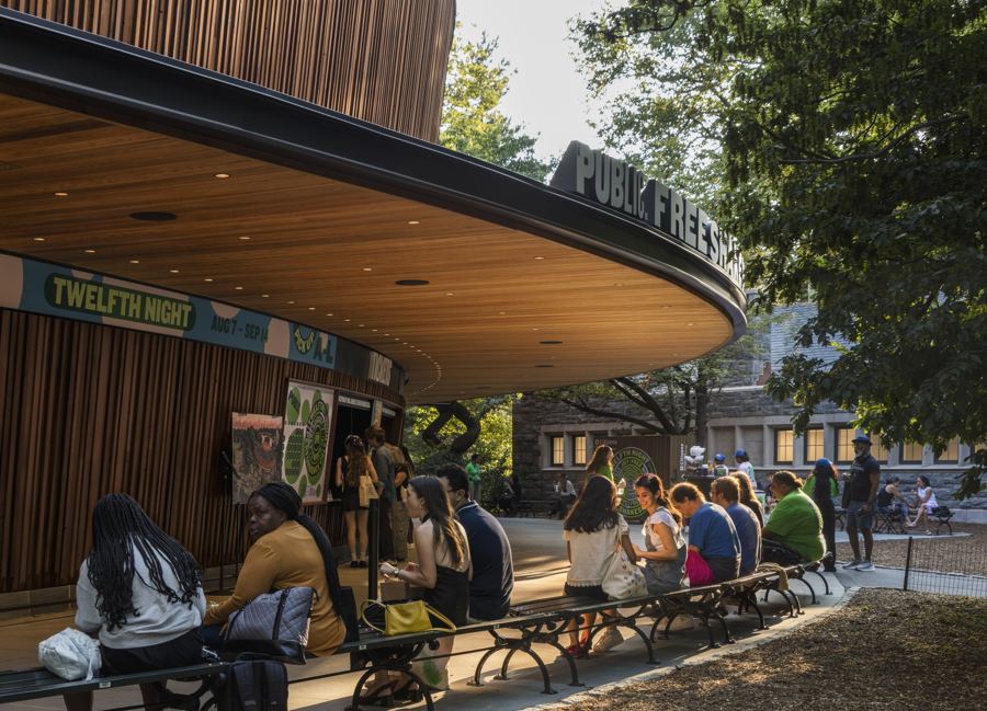 Theatergoers on benches beneath the Delacorte's curved wooden canopy, a Twelfth Night poster beside the gate