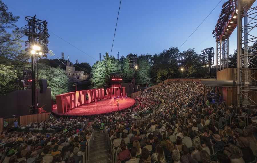 A full house at the Delacorte Theater at dusk, the red stage lit for Twelfth Night under the light towers