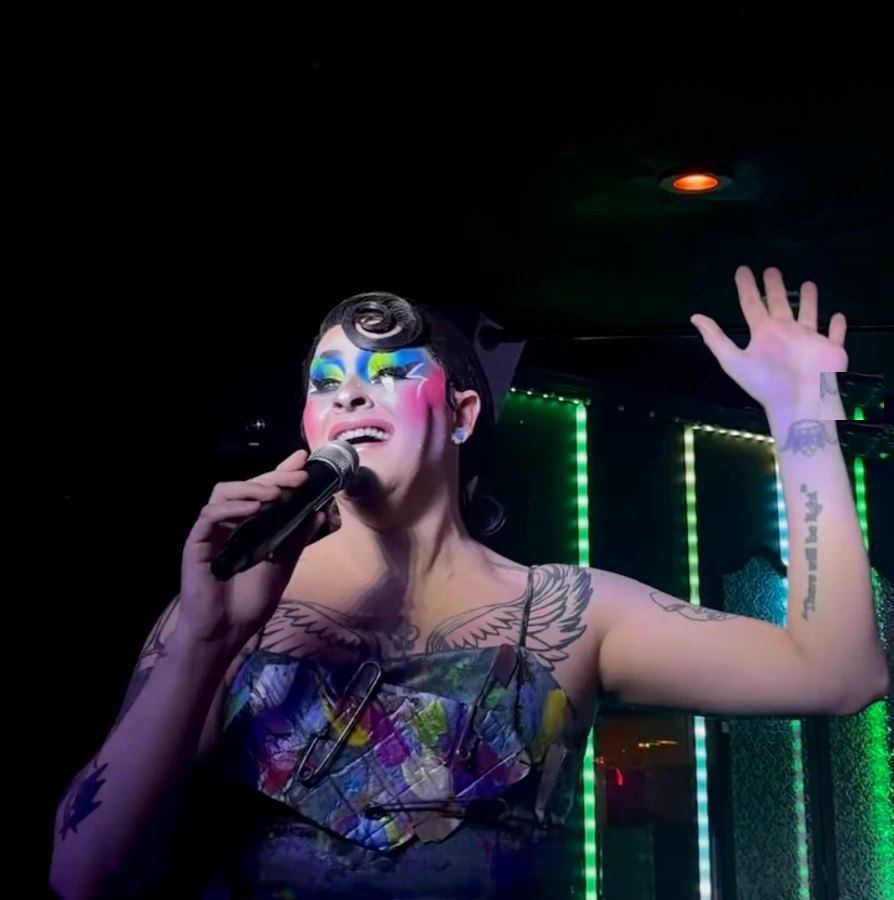 A performer in bright makeup singing under the green-lit panels at The Duplex on Christopher Street