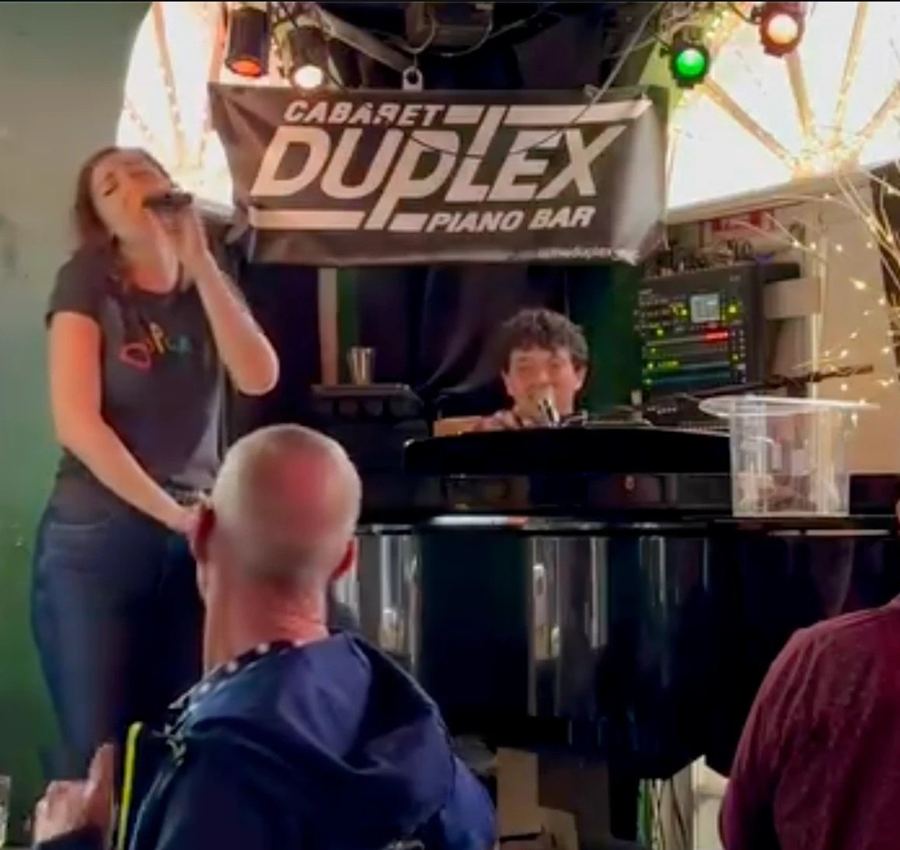 A singer and the pianist at the grand piano under the 'Cabaret Duplex Piano Bar' banner, regulars gathered round