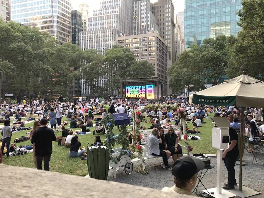 Crowds settling on the Bryant Park lawn on a summer evening before a Movie Night, with Midtown towers behind