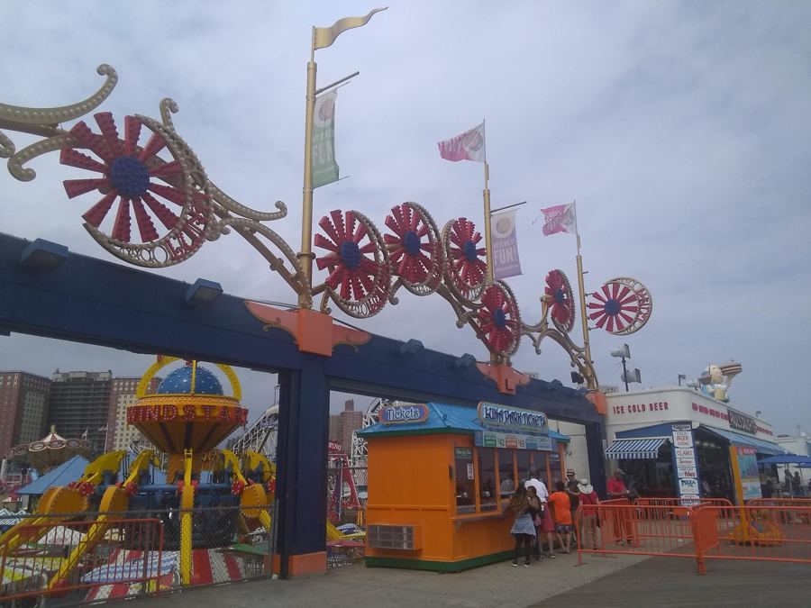 The red pinwheel arches over the boardwalk entrance to Luna Park in Coney Island, with colorful kiddie rides and ticket booths behind