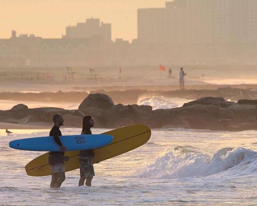 Two surfers carrying boards out of the morning surf at Rockaway Beach, with apartment towers hazy behind