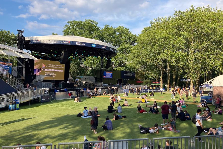 The SummerStage band shell and lawn in Central Park on a summer afternoon, with a small crowd on the grass