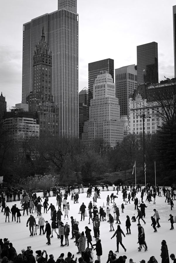 Black-and-white view of skaters on Wollman Rink in Central Park, with the Midtown skyline including the Sherry-Netherland rising behind the trees