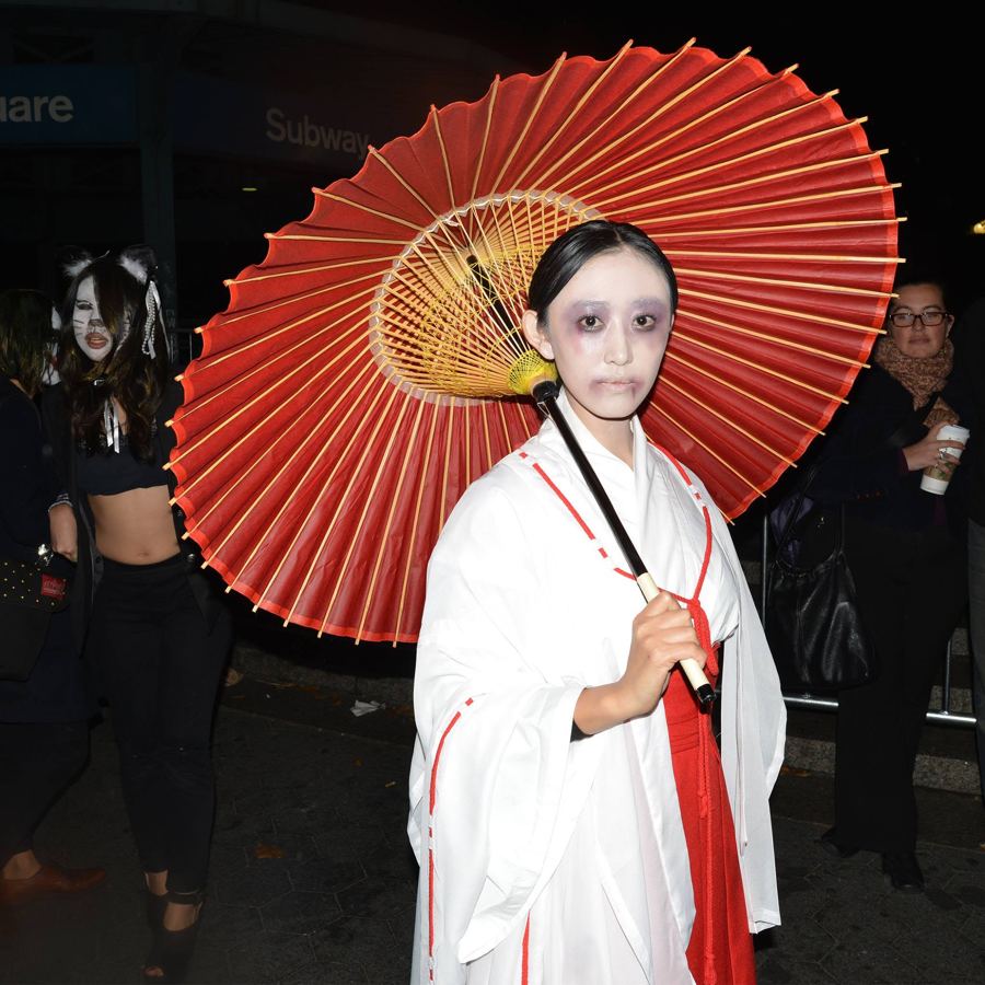 A woman in a white-and-red costume with pale makeup and a red paper umbrella by the Union Square subway entrance on Halloween night