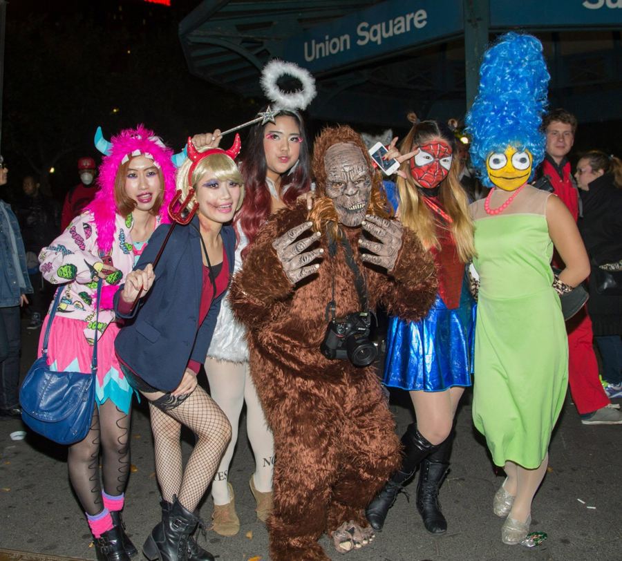 A costumed group, with an angel, a devil, Marge Simpson and a furry beast, under the Union Square subway sign on Halloween night