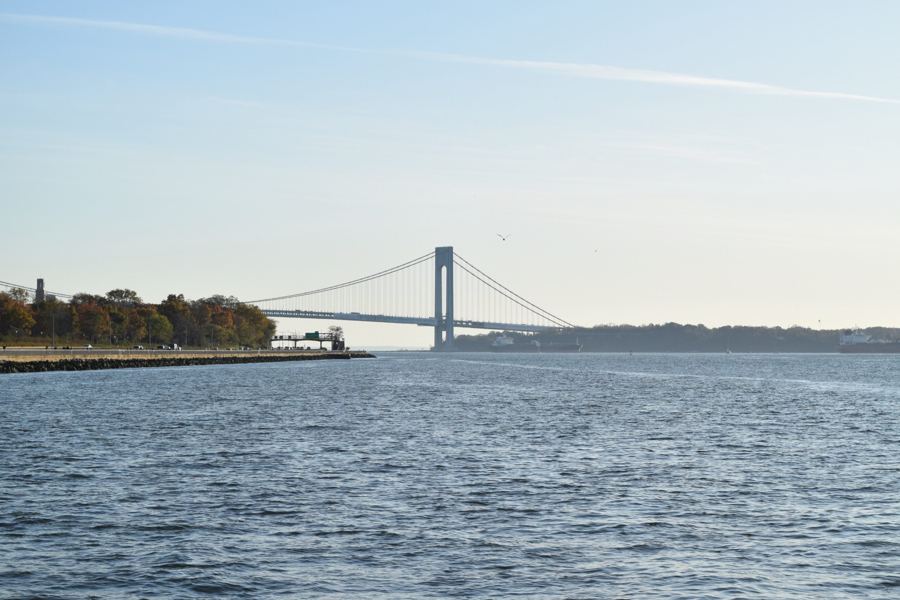 The Verrazzano-Narrows Bridge stretching across the Narrows beyond the Shore Road shoreline, seen over open water from the 69th Street pier in Bay Ridge.