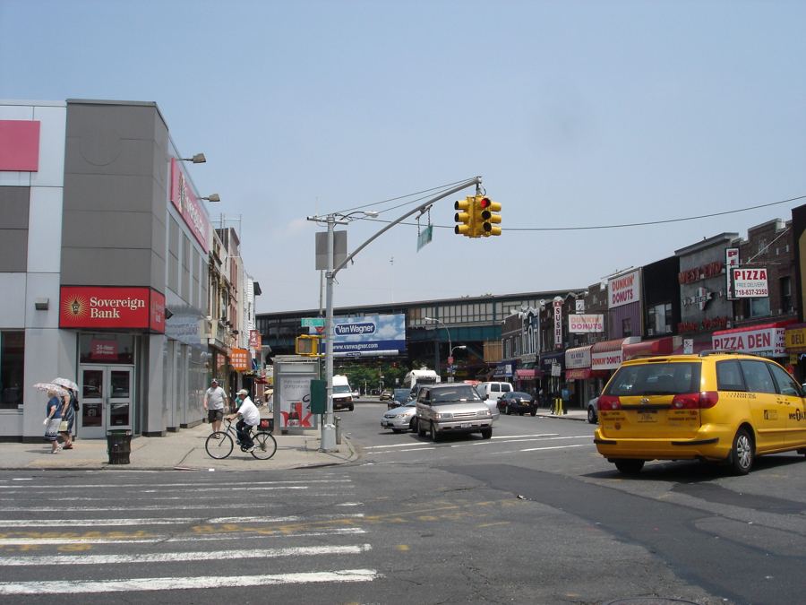 Looking along 18th Avenue in Bensonhurst toward the elevated D train over 86th Street, with storefronts, a cyclist and a yellow taxi.