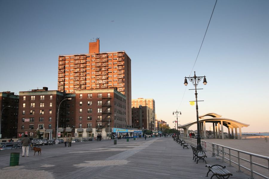 The wide wooden Riegelmann Boardwalk at Brighton Beach in warm evening light, with brick apartment buildings on one side and a beach pavilion and lamp posts on the other.