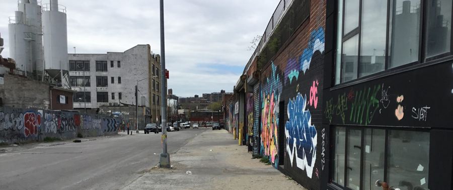 A Bushwick street lined with graffiti-covered warehouse walls, with industrial silos rising behind