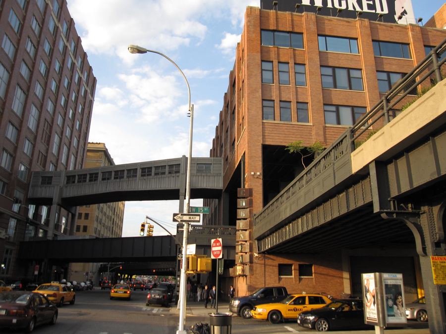 The High Line's elevated rail viaduct and skybridges passing through the brick Chelsea Market complex at 10th Avenue and 15th Street under a blue sky.