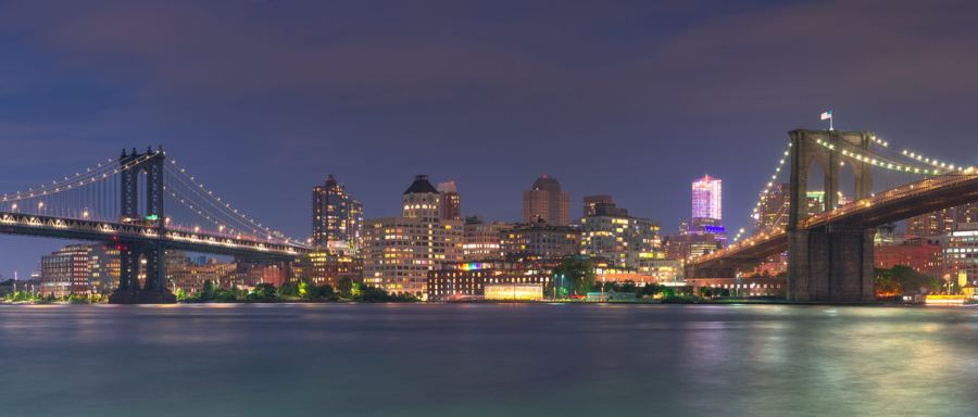 Dumbo lit up at night between the Manhattan and Brooklyn Bridges, seen across the East River