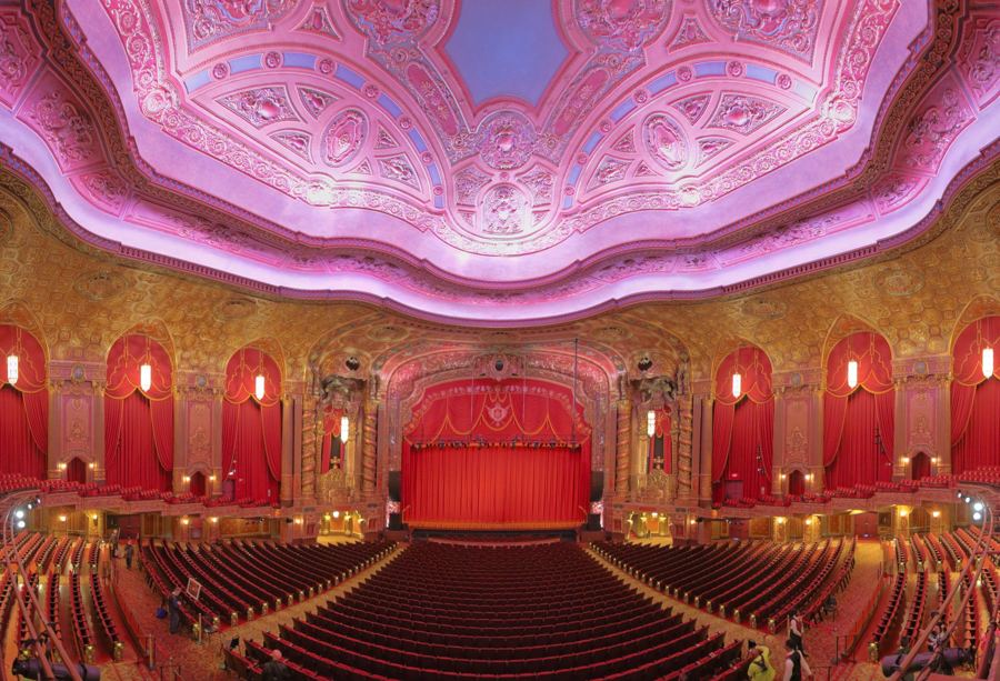 The ornate gilded auditorium of the Kings Theatre in Flatbush, seen from the balcony toward the red stage curtain under a pink-lit domed ceiling.