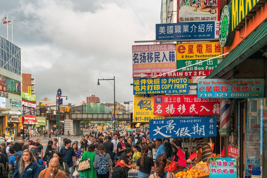 Crowded Main Street in Flushing with stacked bilingual storefront signs and produce stands at the curb