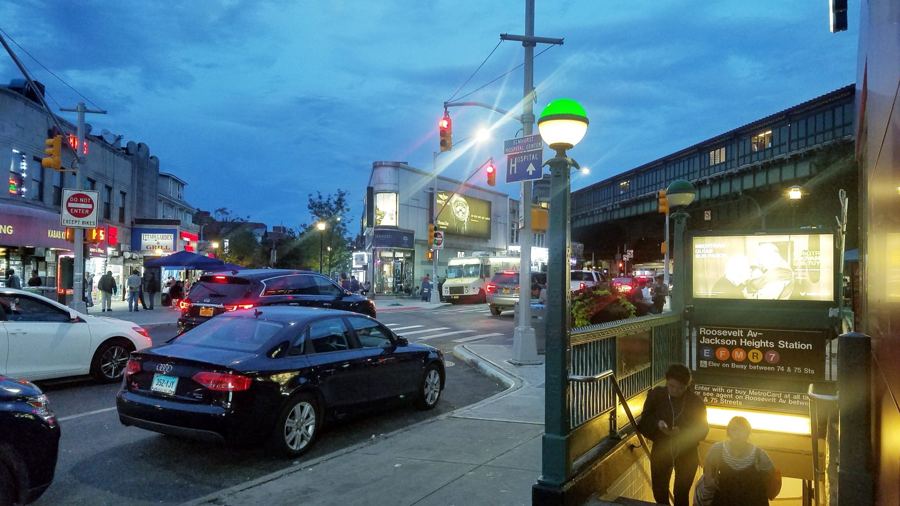 Roosevelt Avenue in Jackson Heights at night, storefront signs and food carts glowing under the elevated 7 train