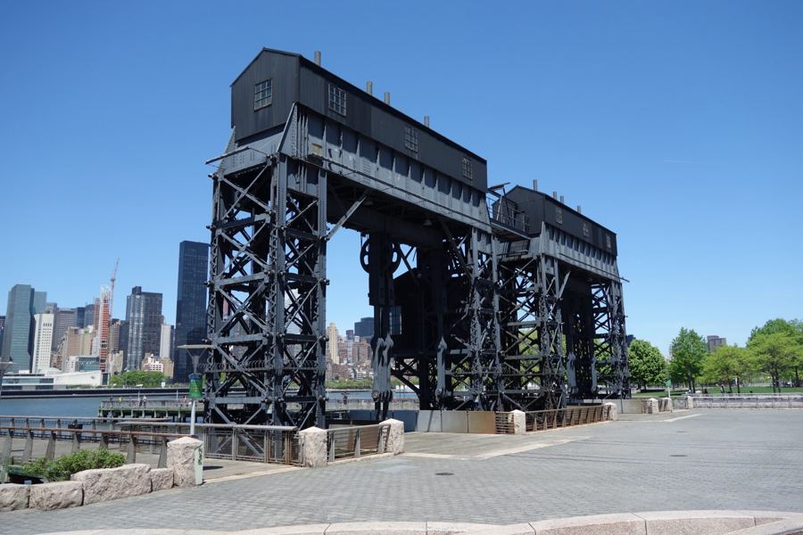 Two black steel railroad gantries stand on the paved waterfront plaza of Gantry Plaza State Park under a clear blue sky, with Midtown Manhattan's towers across the East River.