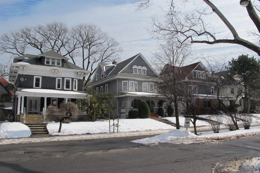 Shingled Victorian houses with deep porches lining a wide, tree-lined Midwood street after a light snowfall.
