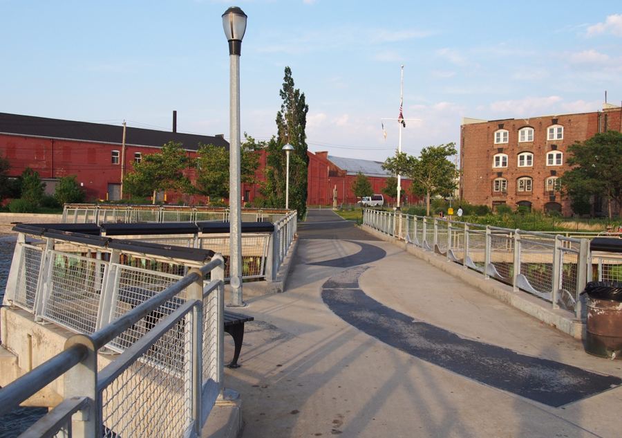 The railed walkway of Valentino Pier leading back toward Red Hook's red-brick waterfront warehouses in warm late-afternoon light.