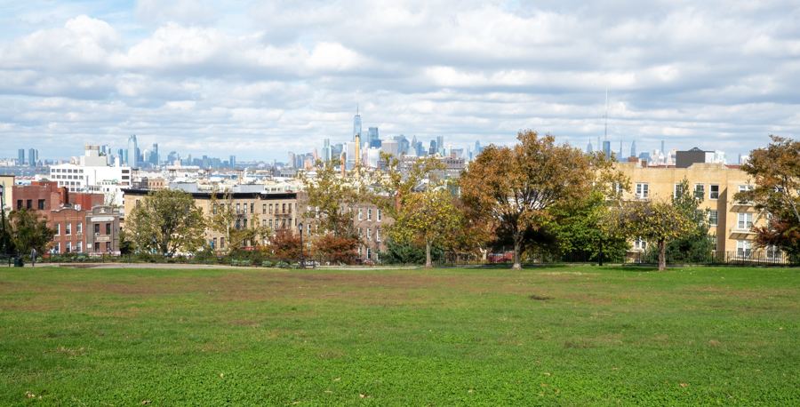 The lower Manhattan skyline seen across the rooftops of Sunset Park, Brooklyn, from the park's broad hilltop lawn ringed with autumn trees.