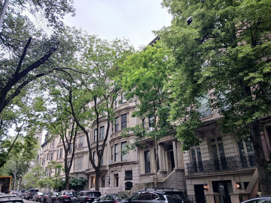 A leafy West 76th Street block of ornate limestone and brownstone rowhouses beneath tall street trees on the Upper West Side.