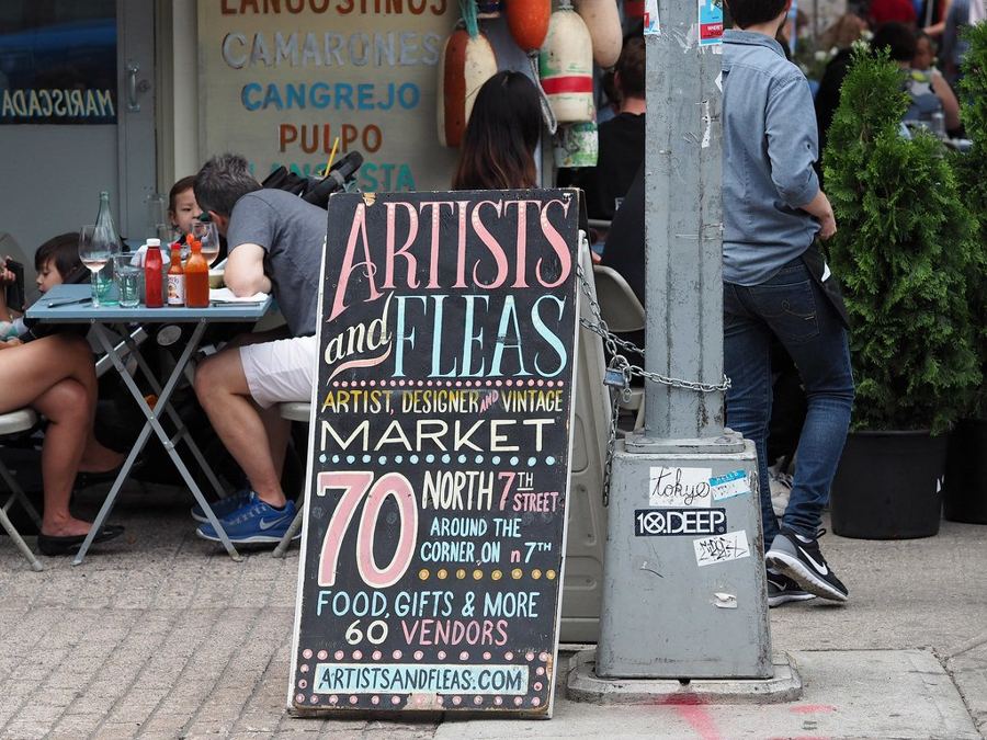 The Artists & Fleas chalkboard sign on the sidewalk at 70 North 7th Street, Williamsburg, pointing shoppers to 60 vendors