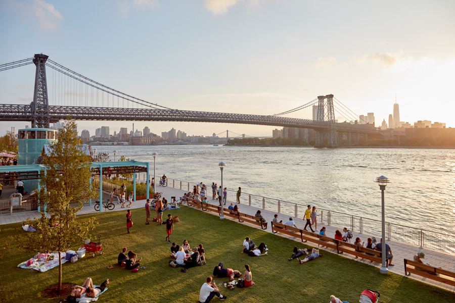 Picnickers on the lawn at Domino Park, with the Williamsburg Bridge and the Manhattan skyline across the East River