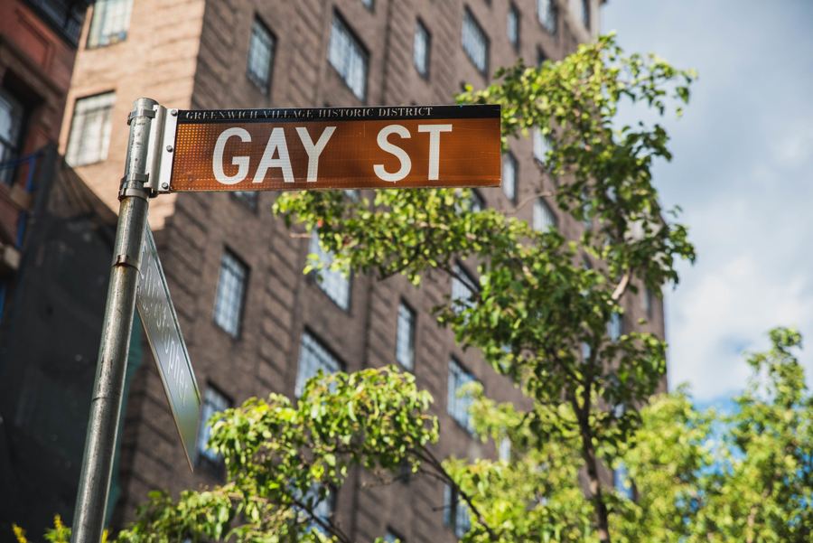 The brown Gay St sign of the Greenwich Village Historic District against brick and summer trees